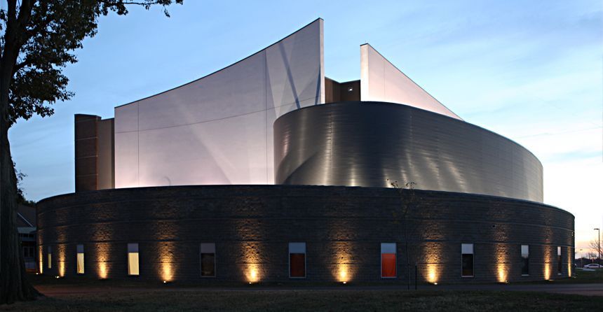 A modern building with a curved stone base and a prominent, angular white facade, illuminated at dusk.