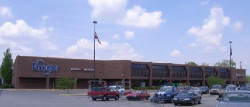A brown, single-story Kroger grocery store with a large parking lot in front under a blue sky with scattered clouds.