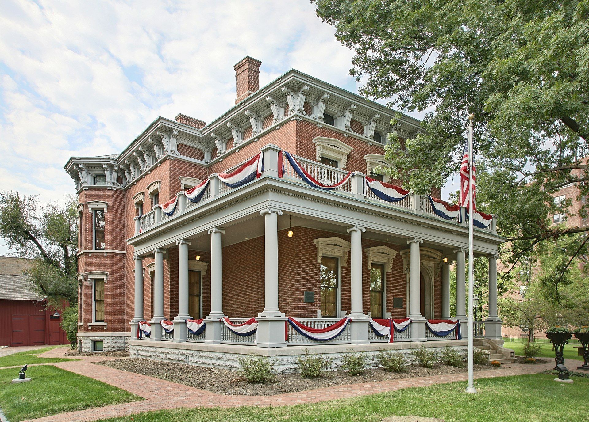 A two-story red brick Italianate house with a wrap-around porch decorated with red, white, and blue bunting.