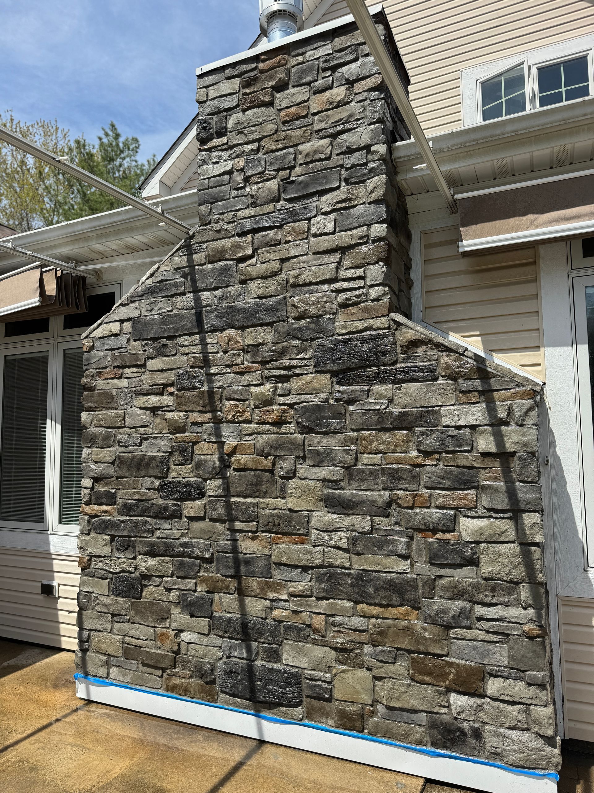 A stone fireplace chimney exterior with multi-colored gray and tan stacked stones against a light siding wall.