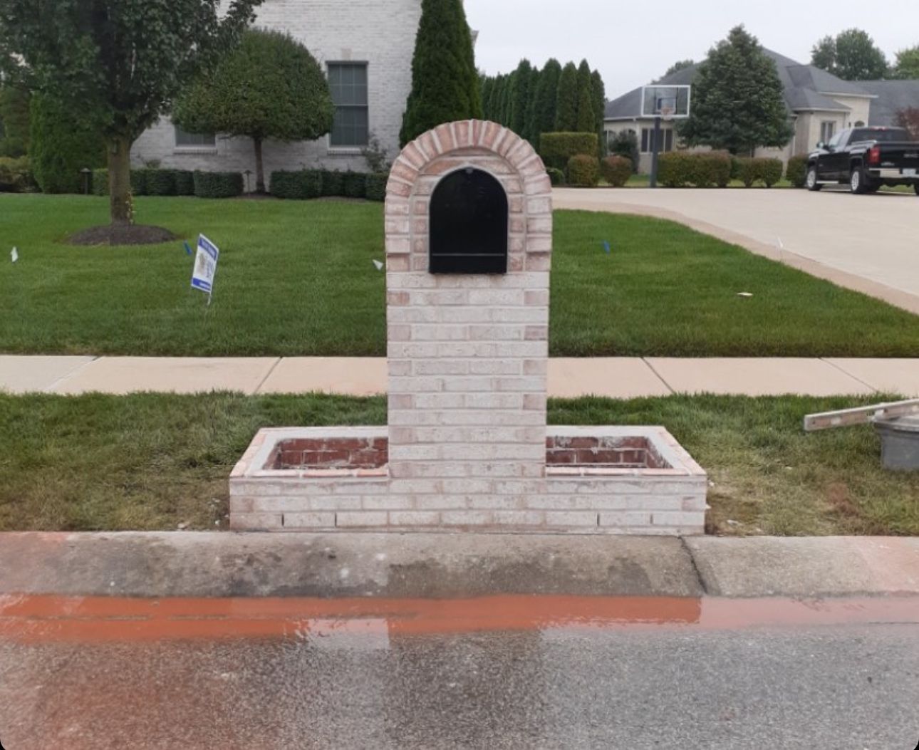 A light-colored brick mailbox structure with a rounded top and built-in side planters sits curbside in a residential yard.