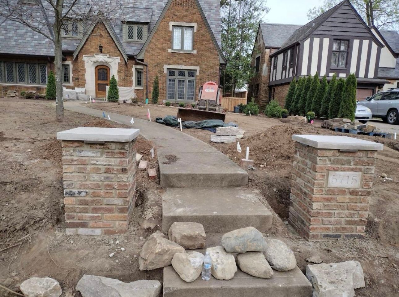 A new concrete walkway with stone steps leads to a brick house under construction, flanked by two unfinished brick pillars.