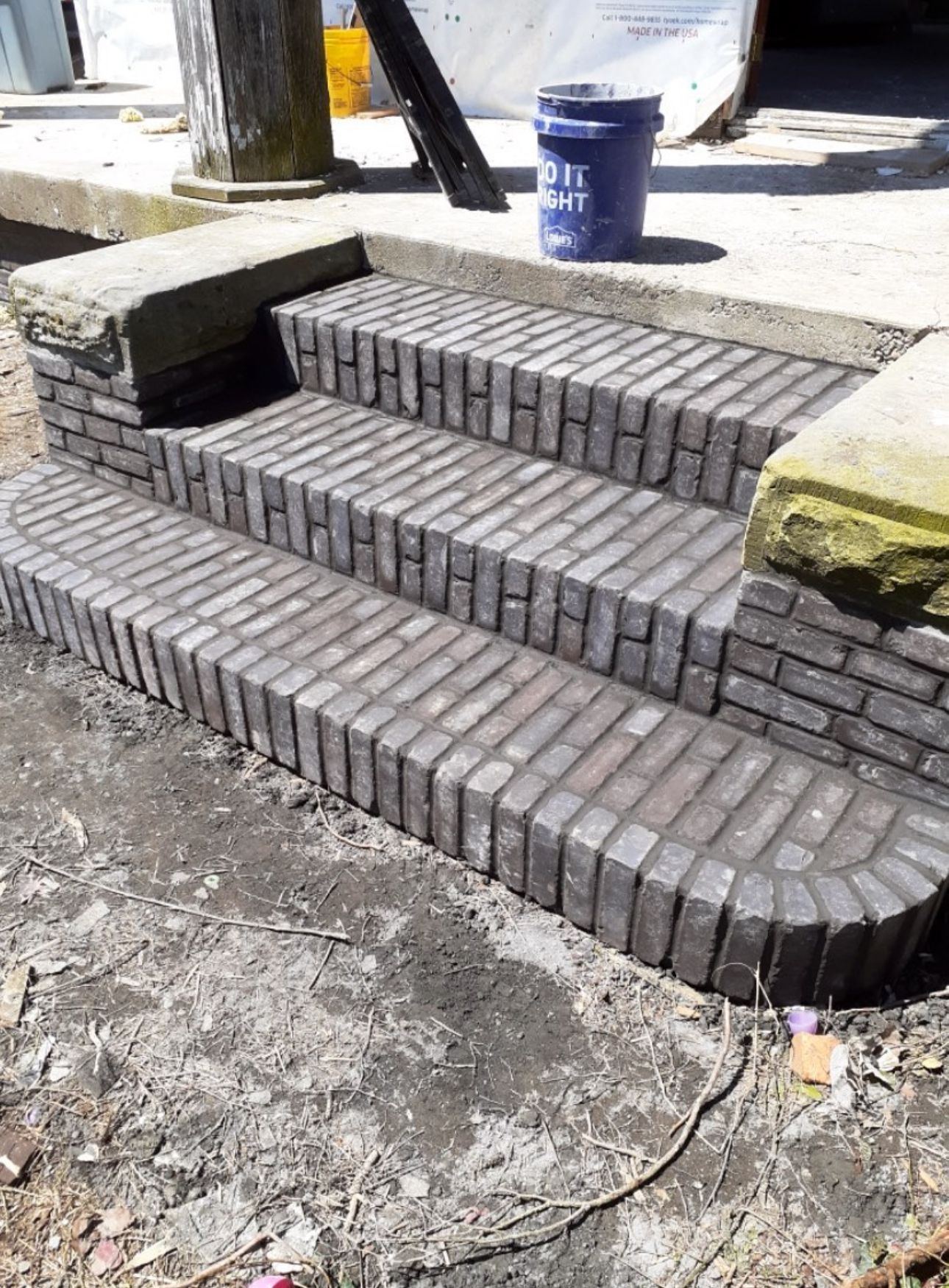 A set of three new brick steps leading up to a porch, flanked by stone blocks, with a blue bucket on the porch above.