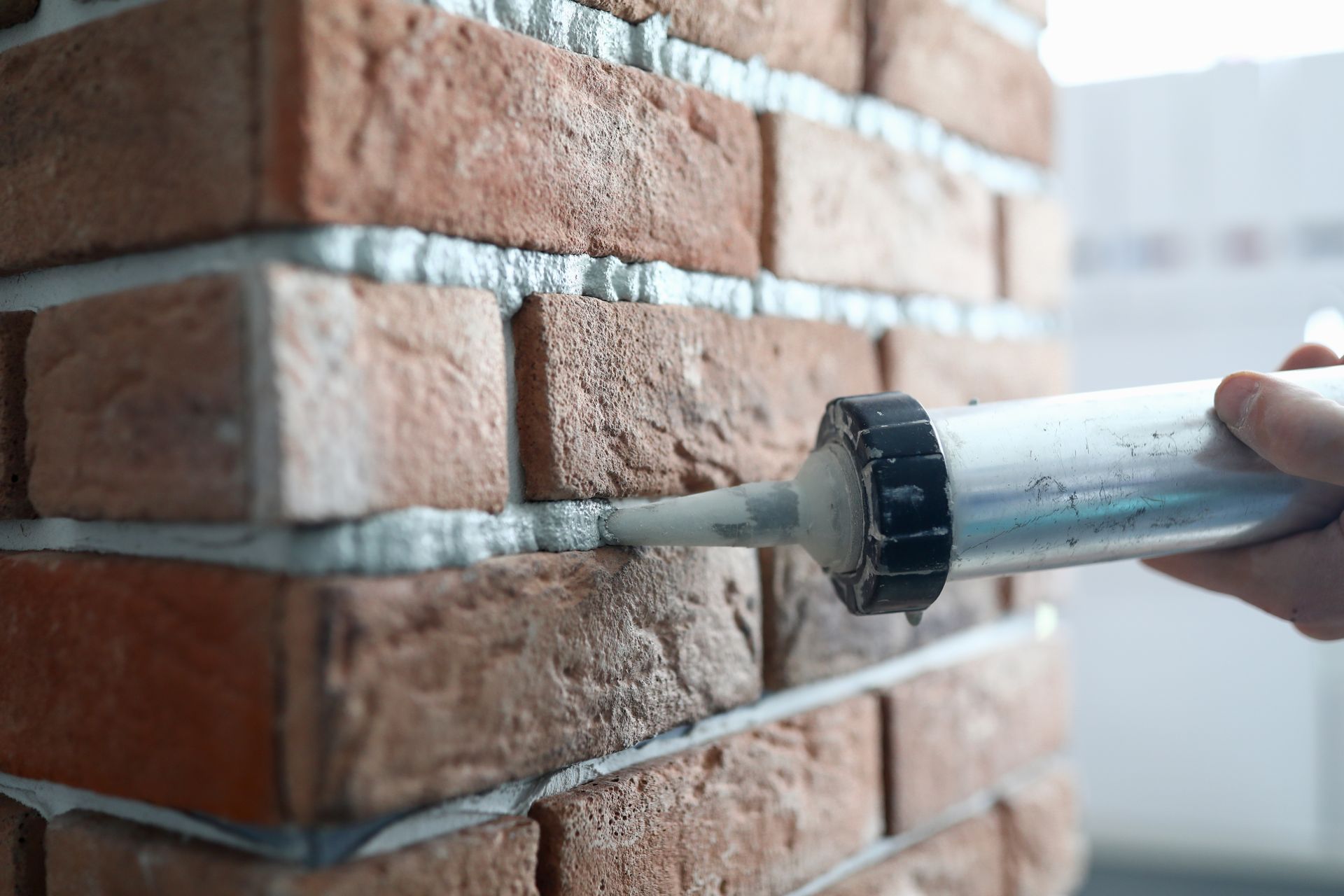 A hand uses a caulking gun to apply mortar or sealant into the gaps between red bricks on a wall.