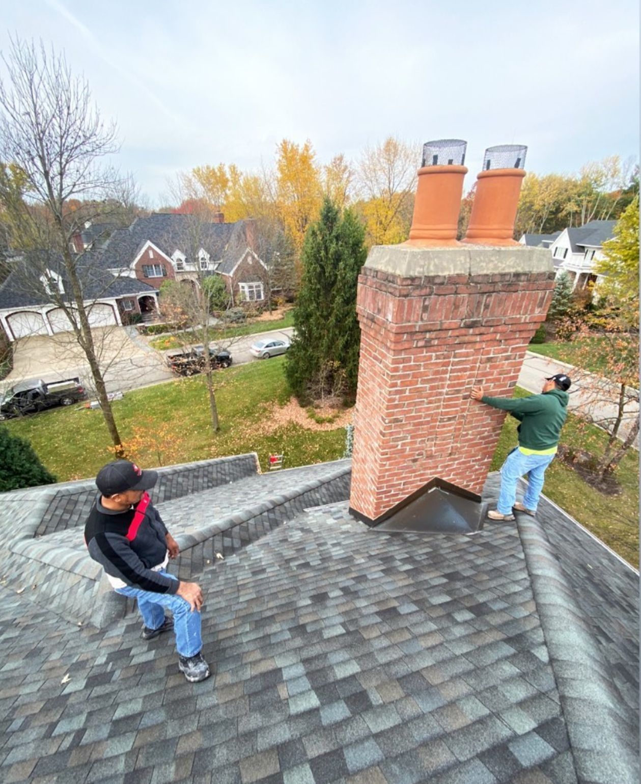 Two workers inspect a brick chimney on a residential roof during a fall day.