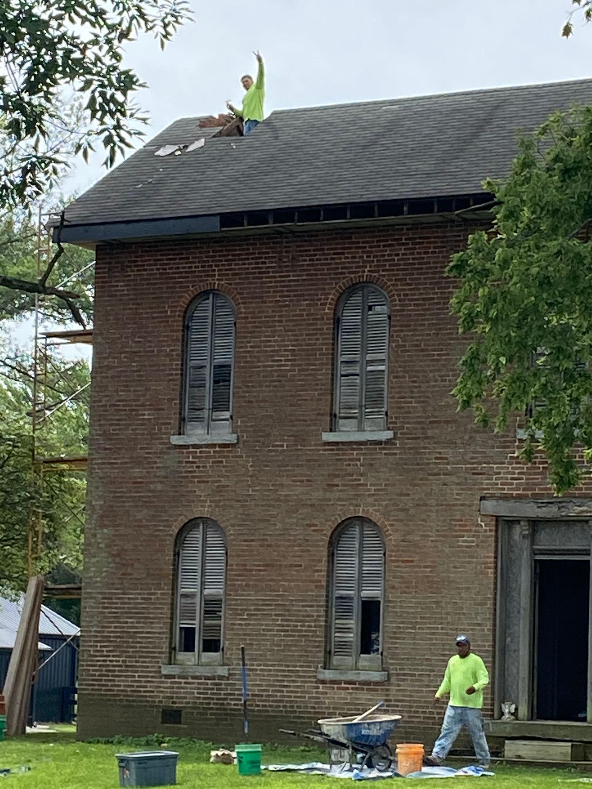 Two workers in high-visibility yellow shirts perform repairs on a brick building, one on the roof and one on the ground.