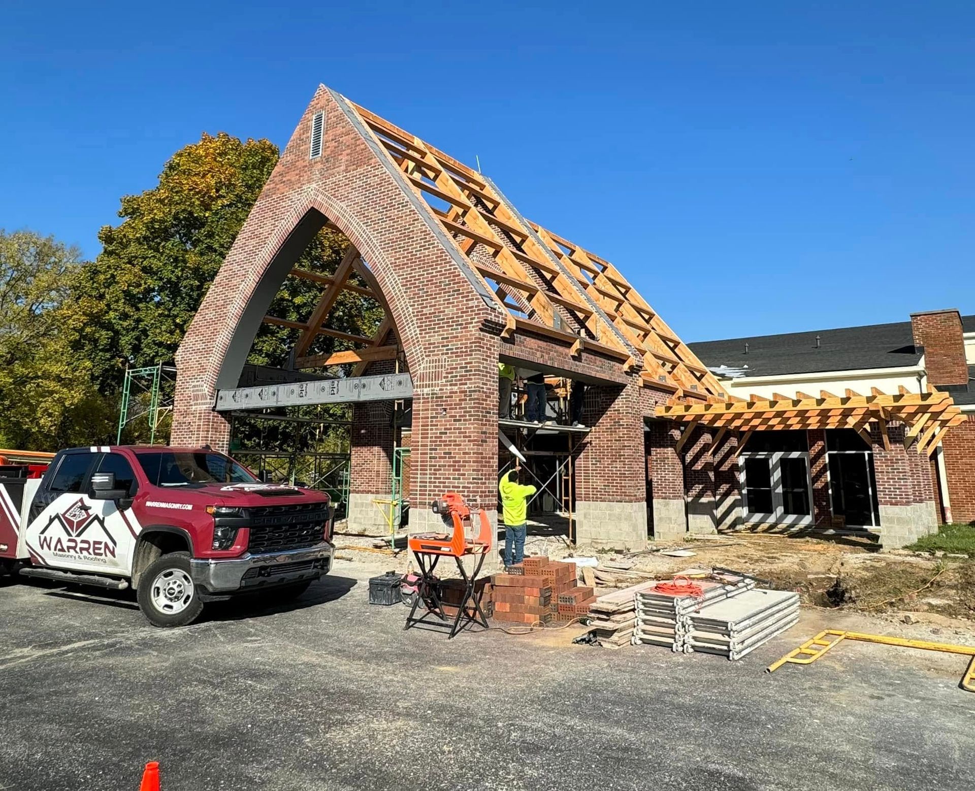 Church roof under construction with exposed wooden trusses, brick facade, and a red pickup in front