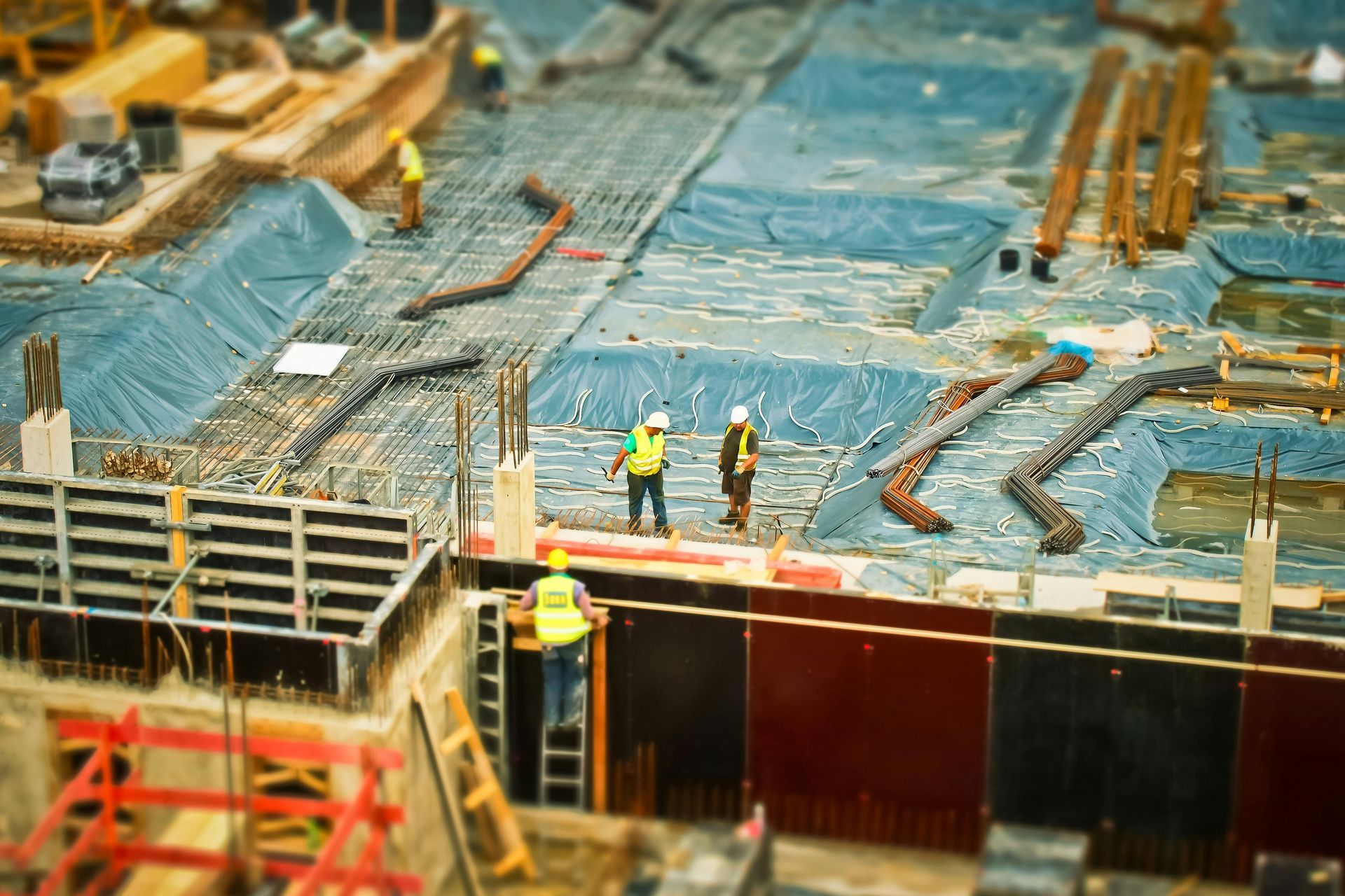 Construction workers in high-visibility vests work on a foundation site with blue coverings and metal structures.