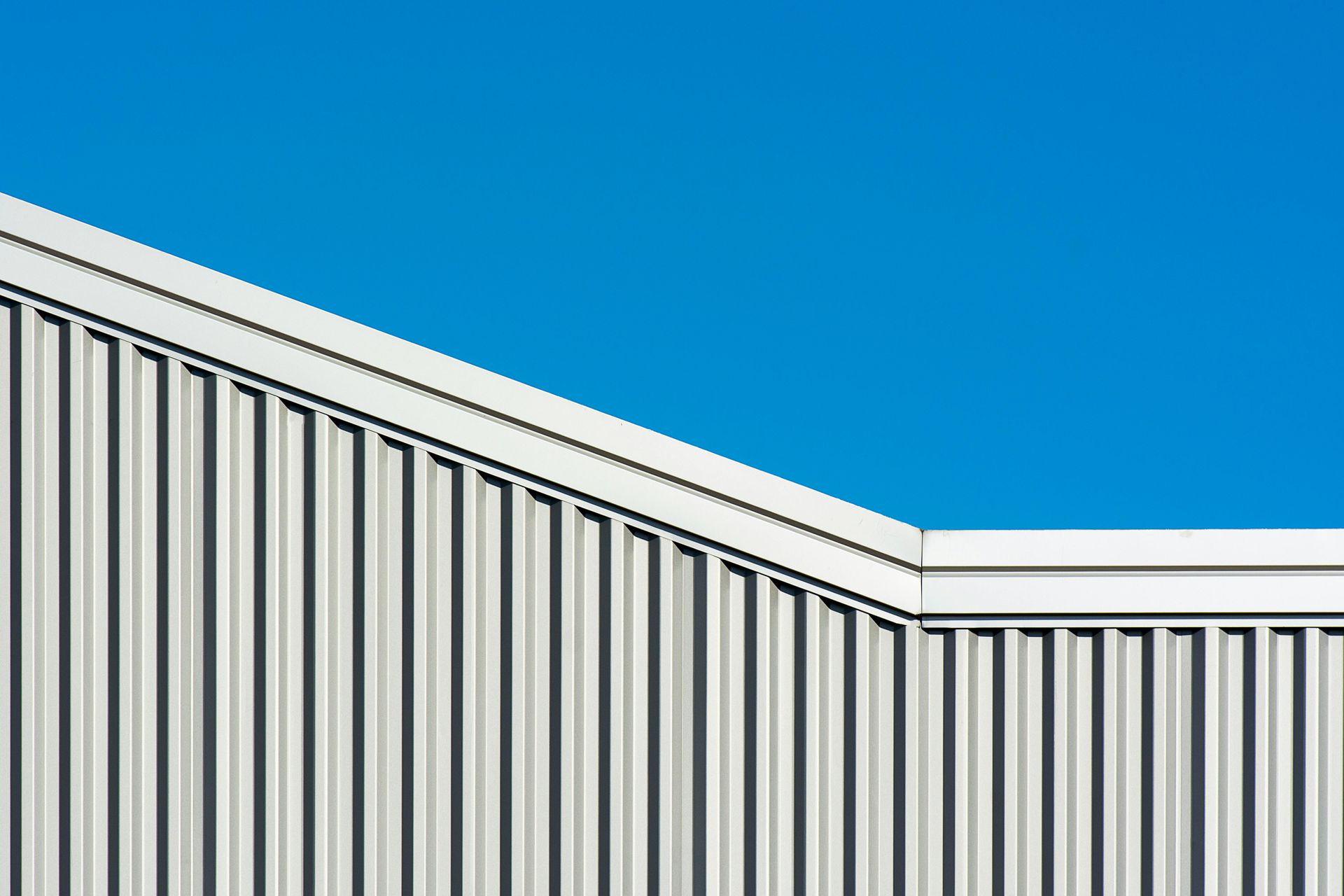 A minimalist view of a light-gray corrugated metal building exterior against a clear, solid blue sky.
