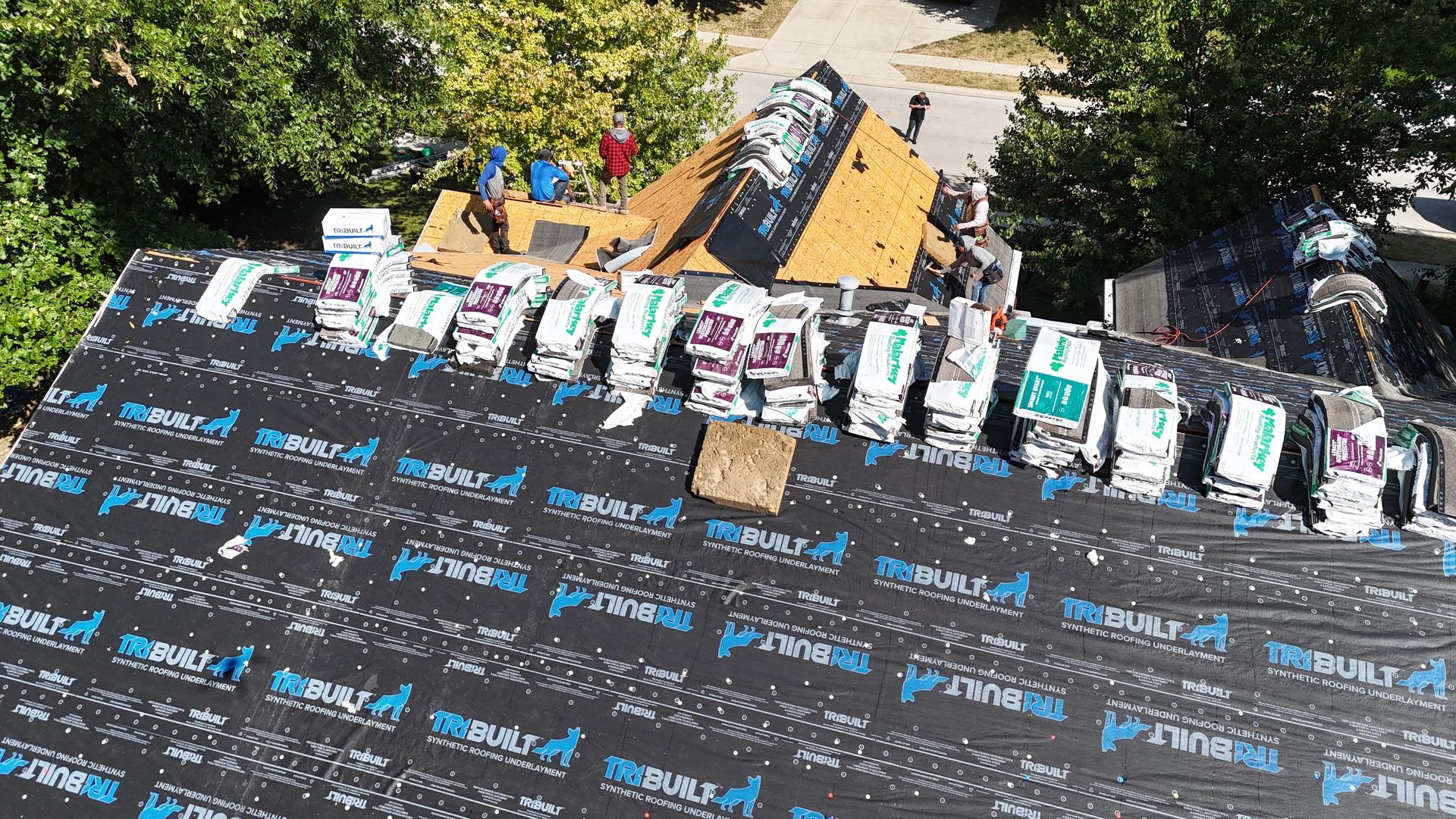Workers installing shingles on a residential roof covered in black synthetic underlayment.