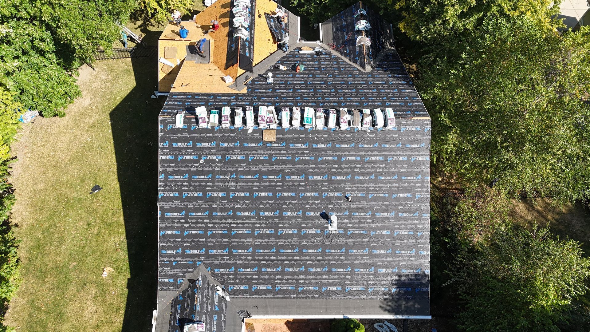 Aerial view of a residential roof under construction, featuring black underlayment, bundled shingles, and exposed decking.