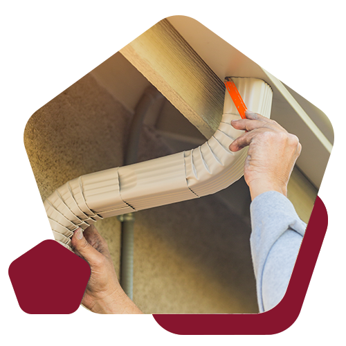 A pair of hands installs a tan, corrugated metal gutter extension along the exterior eaves of a building.