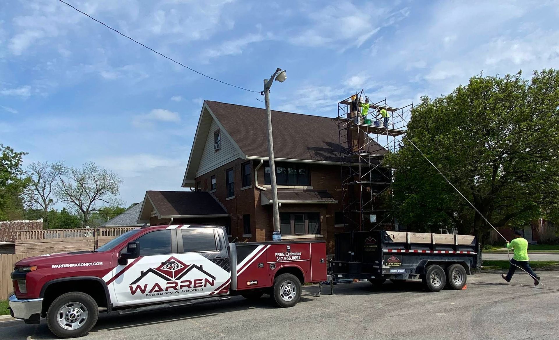 A maroon Warren construction truck pulling a trailer with utility equipment, parked next to a house with workers nearby.