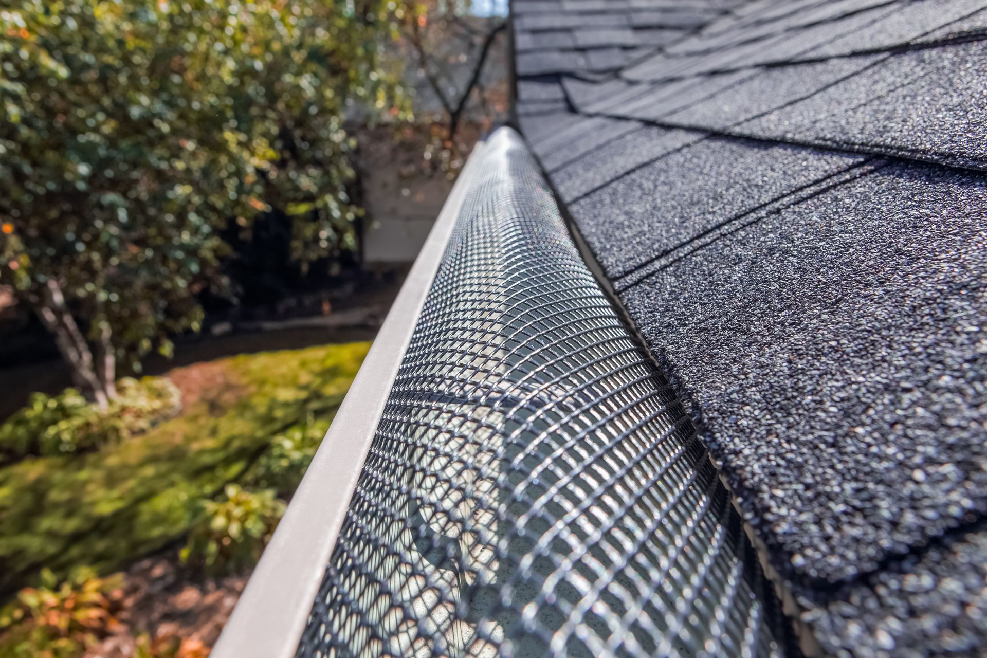 A close-up view of a metal mesh gutter guard installed on a roof with asphalt shingles, overlooking a backyard garden.
