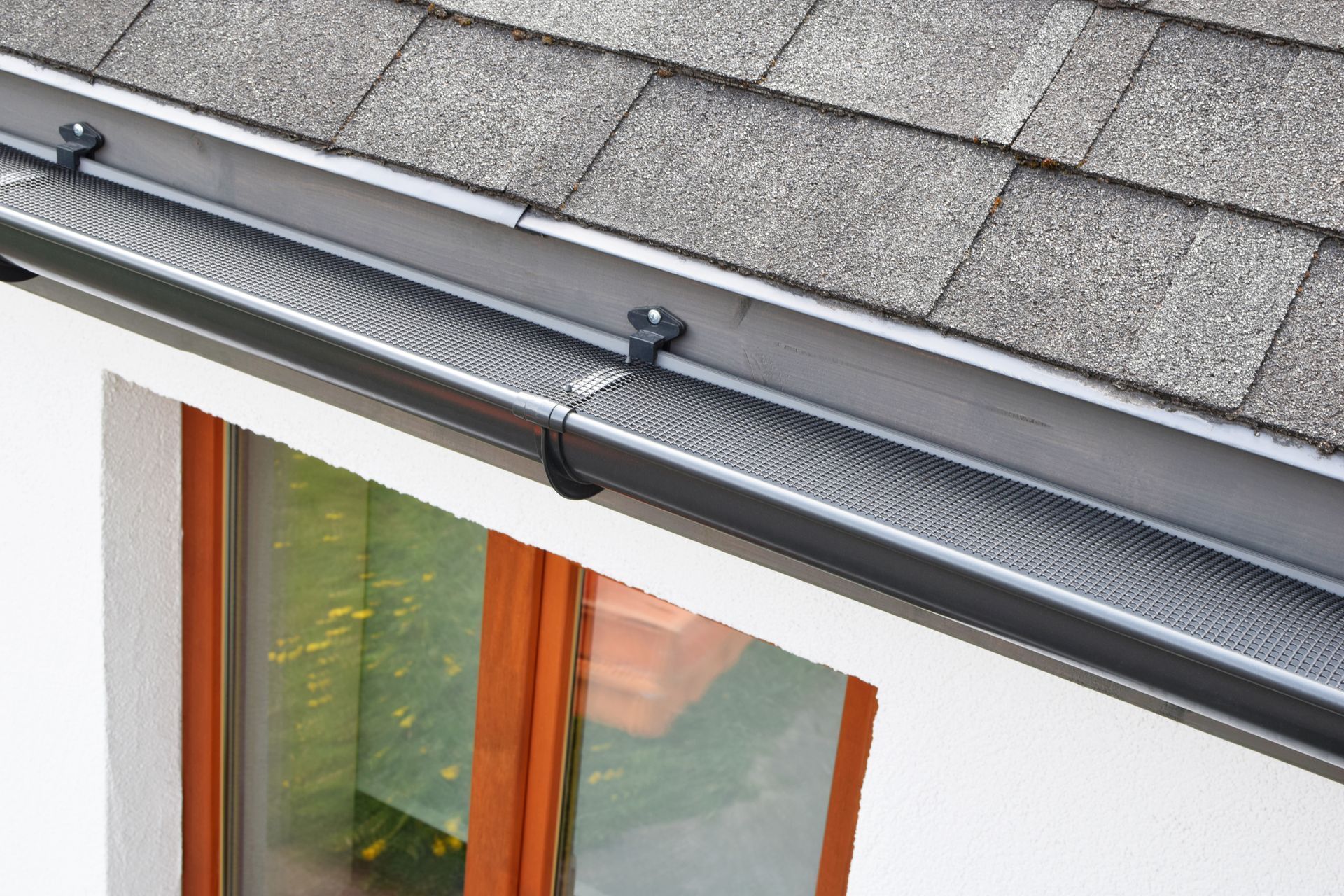 A close-up view of a dark gutter guard installed on a house roof above a window with a white wall frame.