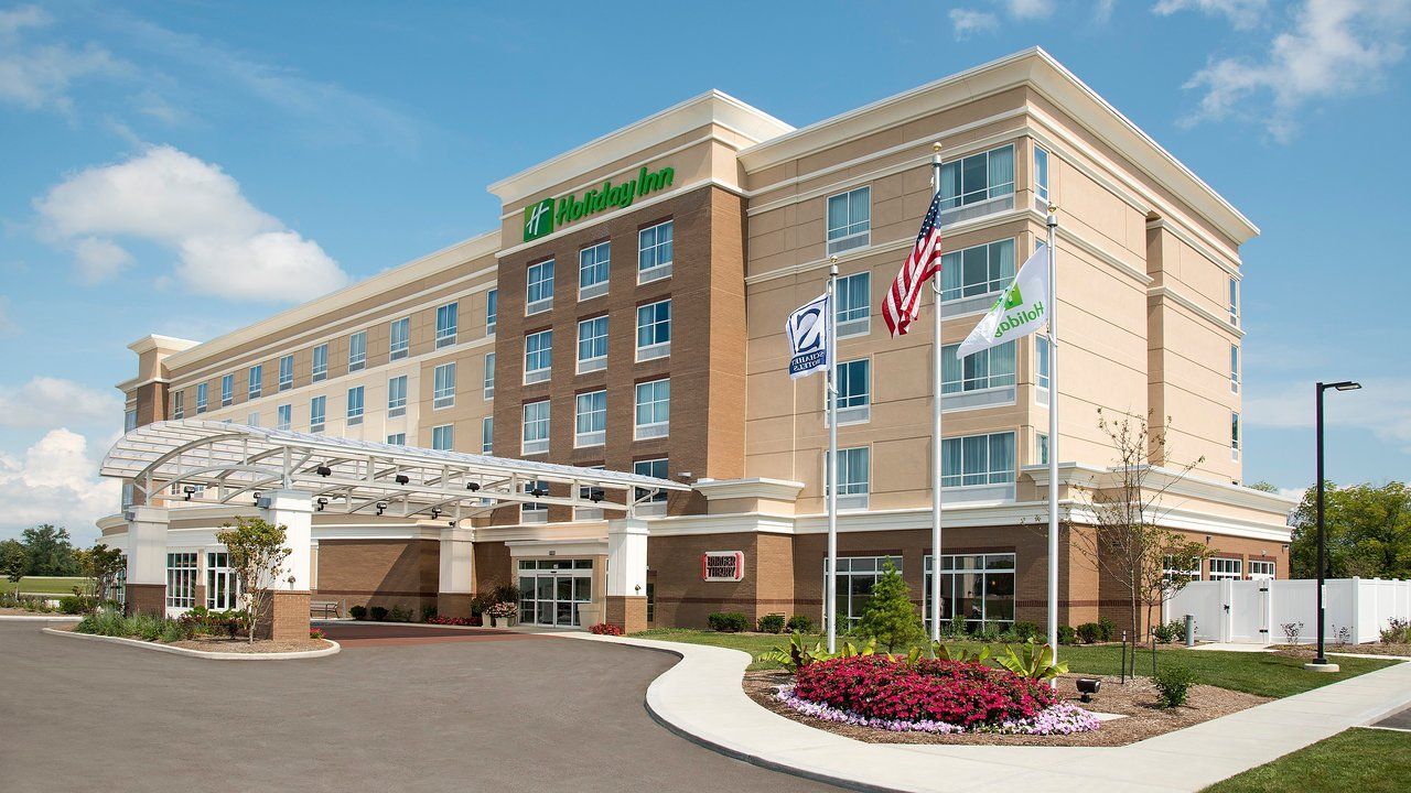 A tan, multi-story Holiday Inn hotel building with a brick facade, covered entrance, and three flags in front.