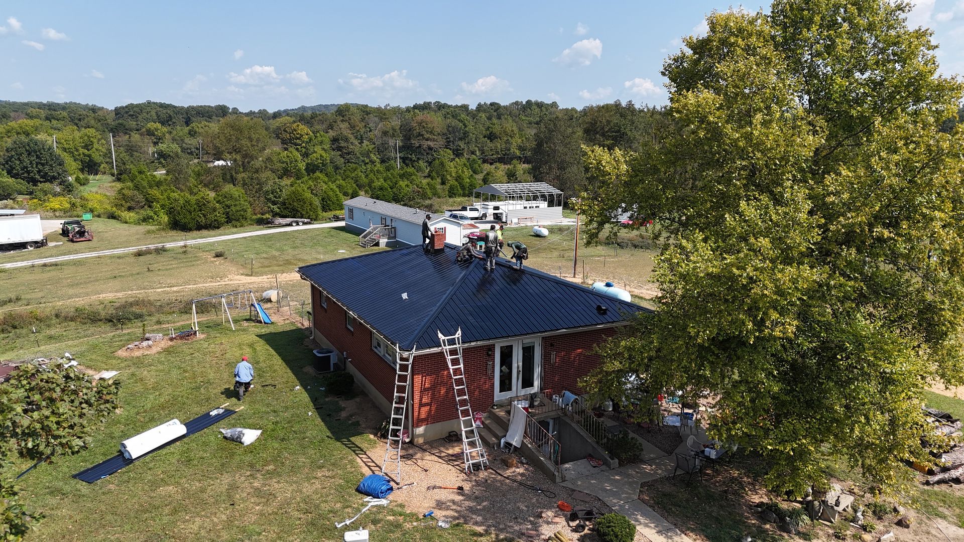 An aerial view shows contractors installing a dark-colored metal roof on a red house surrounded by trees and rural land.
