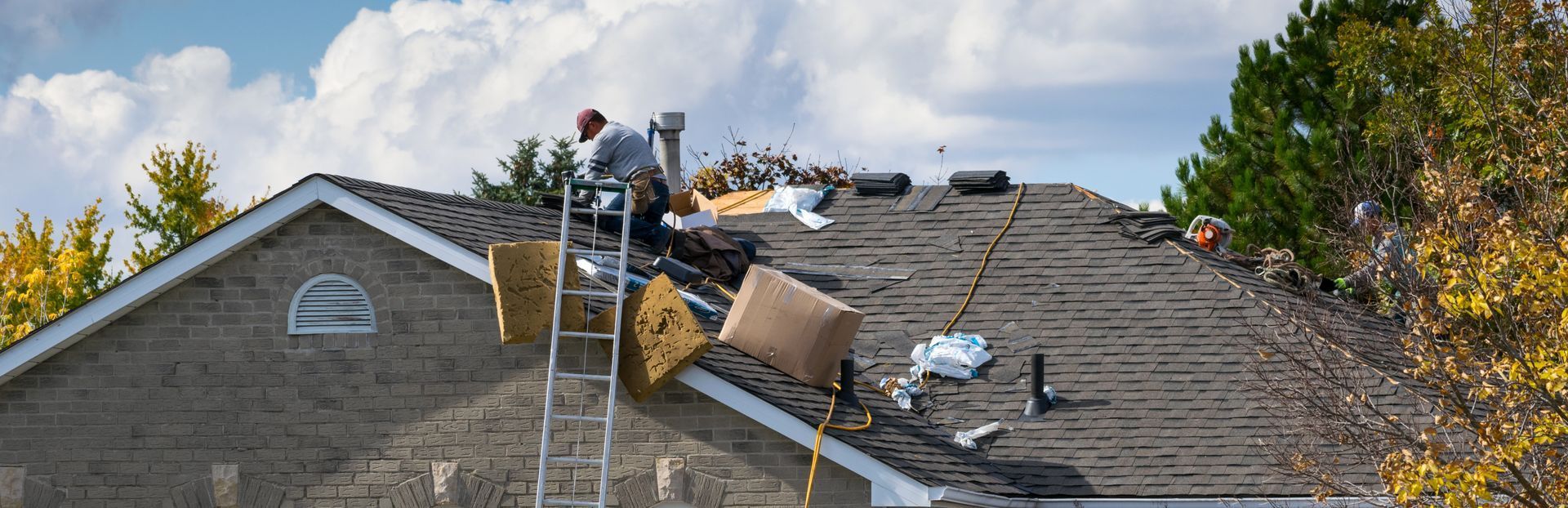 A construction worker installs materials on the sloped shingle roof of a brick house on a sunny day with trees nearby.