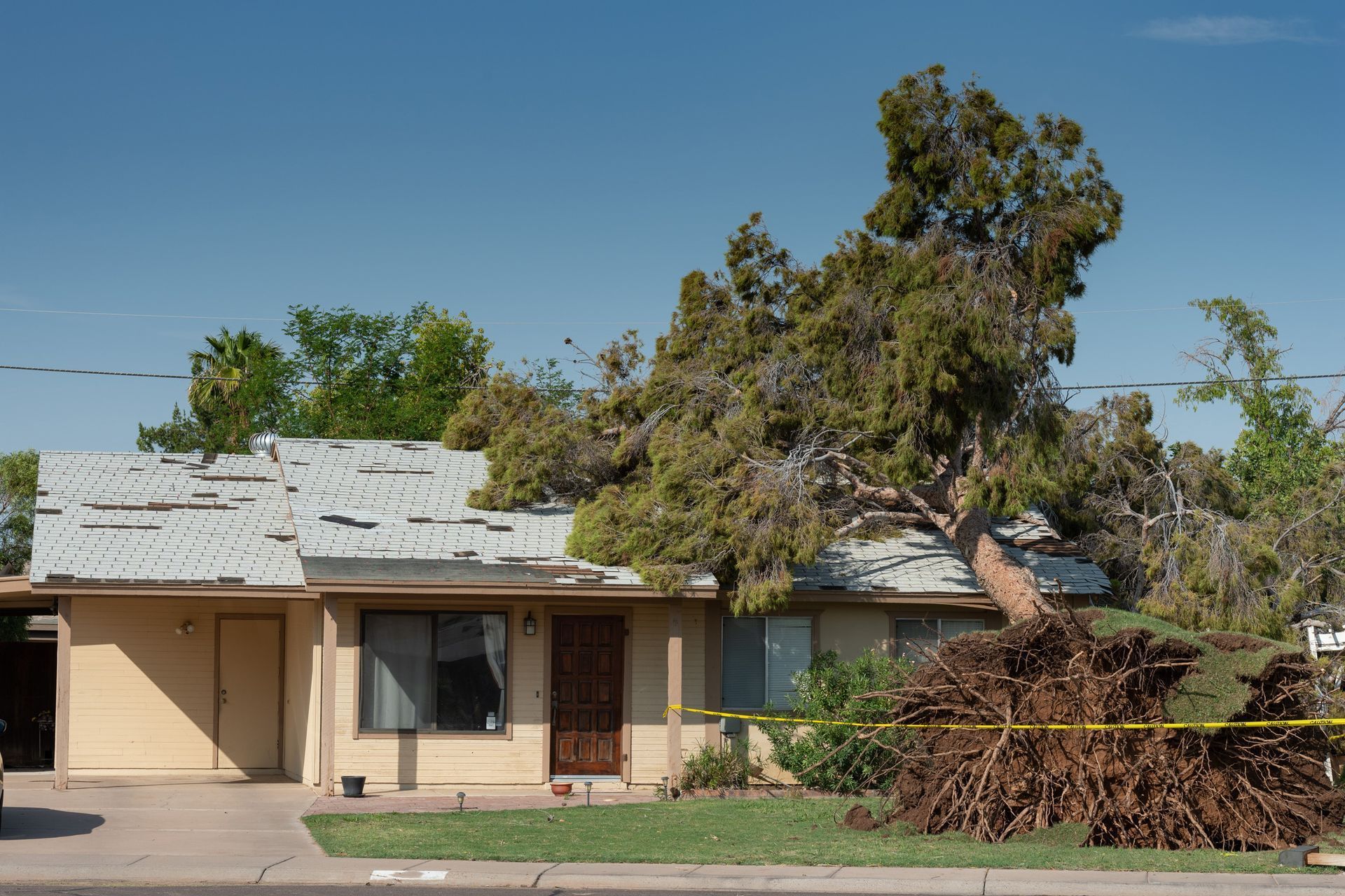 A large tree has fallen onto the roof of a one-story tan house, surrounded by yellow caution tape.