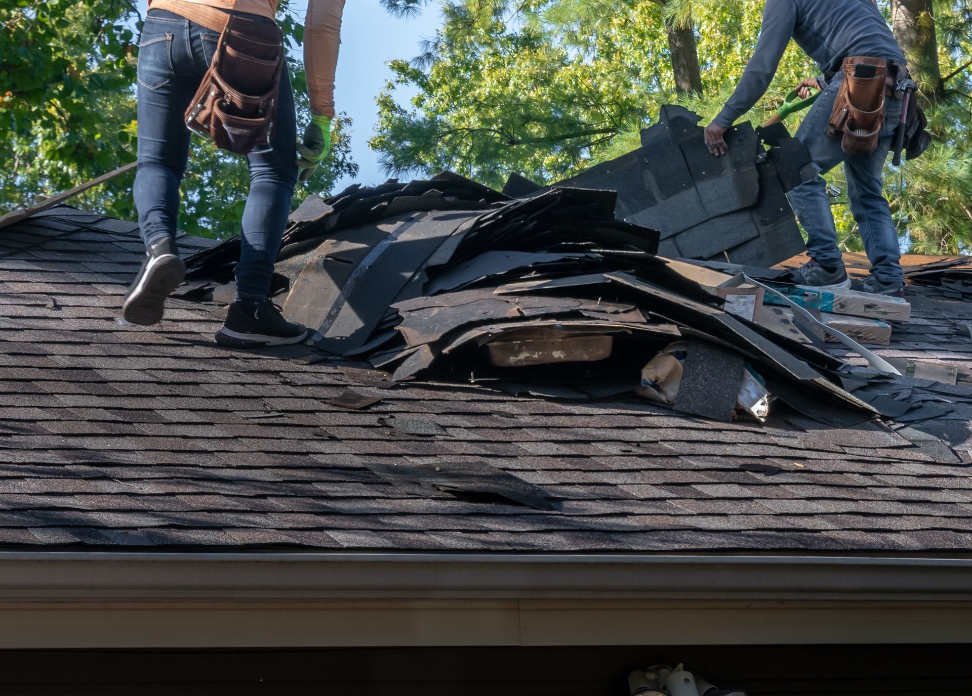 Two workers in blue jeans and tool belts remove old, dark shingles from a sloped roof in Indianapolis.