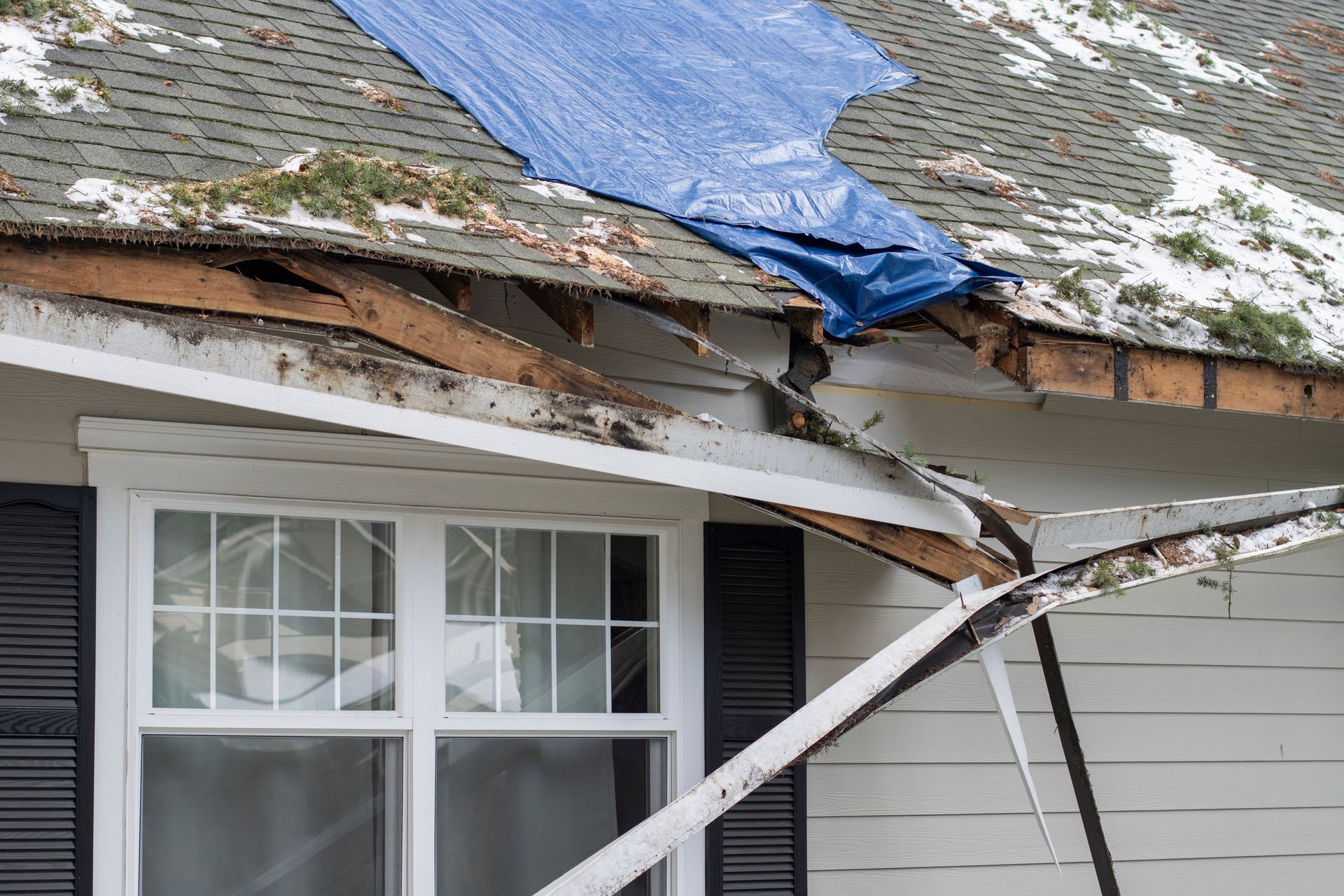 A house exterior with damaged roof eaves, torn wood, and a blue tarp covering part of the shingles.