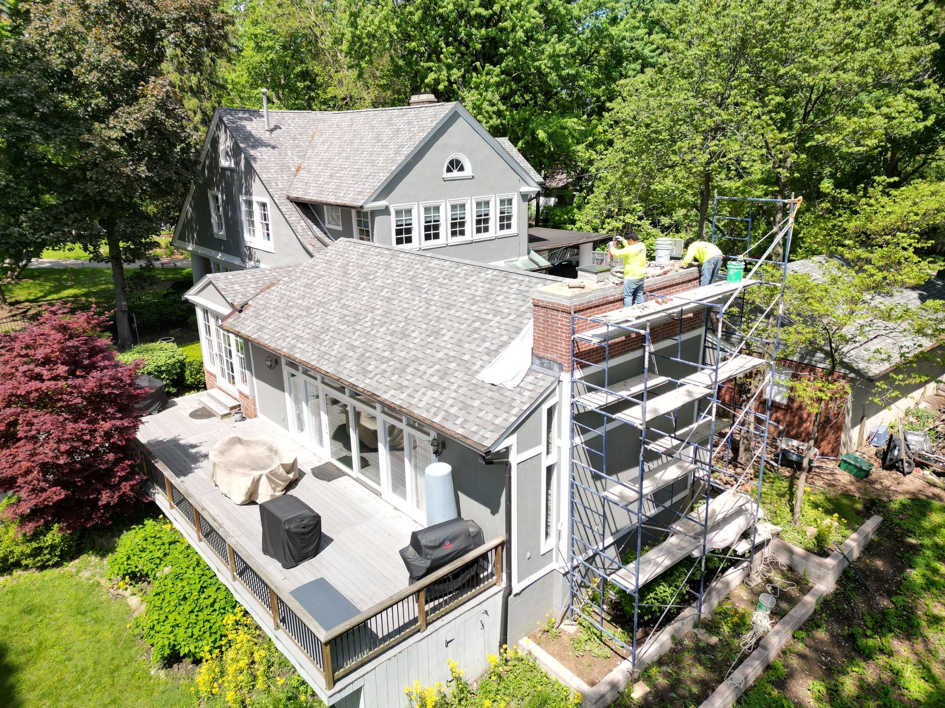 Gray house with rooftop deck and scaffolding, surrounded by trees and a landscaped yard