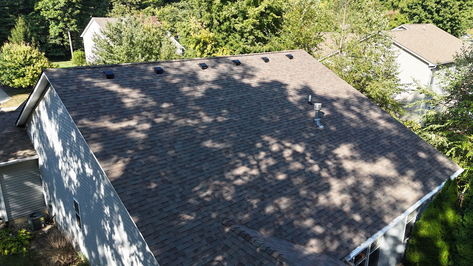 An aerial view of a gray shingled residential roof with several dark vents near the peak, surrounded by green trees.
