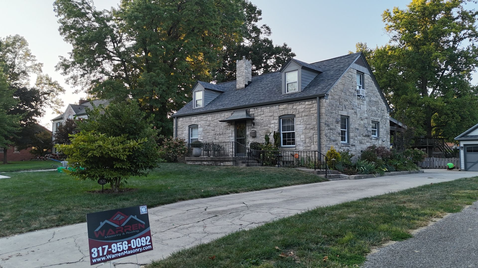 A stone house with a slate roof and dormer windows sits behind a lawn with a real estate sign in the foreground.