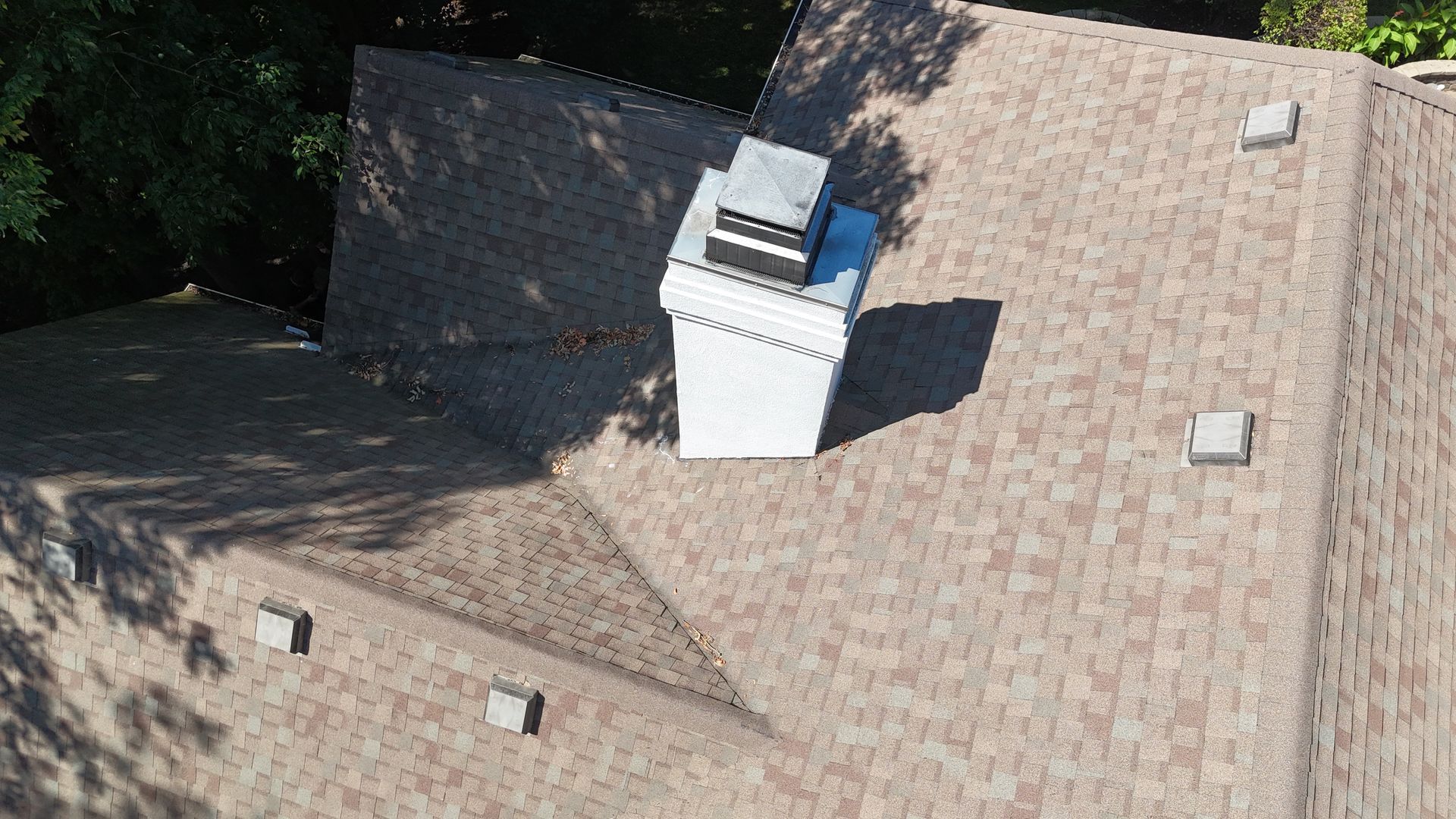 An aerial view of a tan shingled roof featuring a white chimney and several small roof vents surrounded by trees.