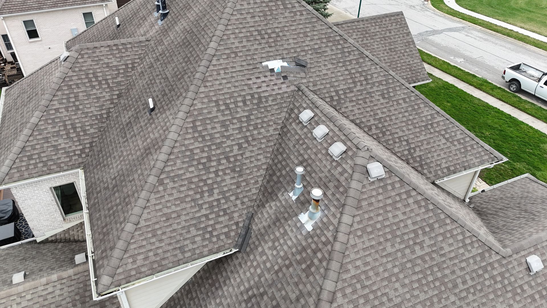 An aerial view of a multi-gabled roof with brown asphalt shingles, roof vents, and plumbing stacks on a residential home.