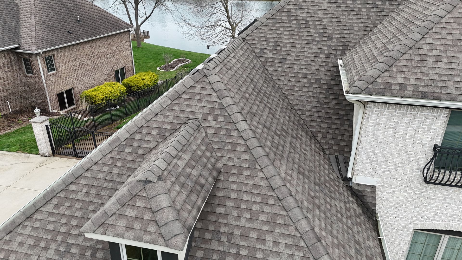 An aerial view of a dark grey shingled roof on a house, featuring a dormer window and a neighboring brick home.