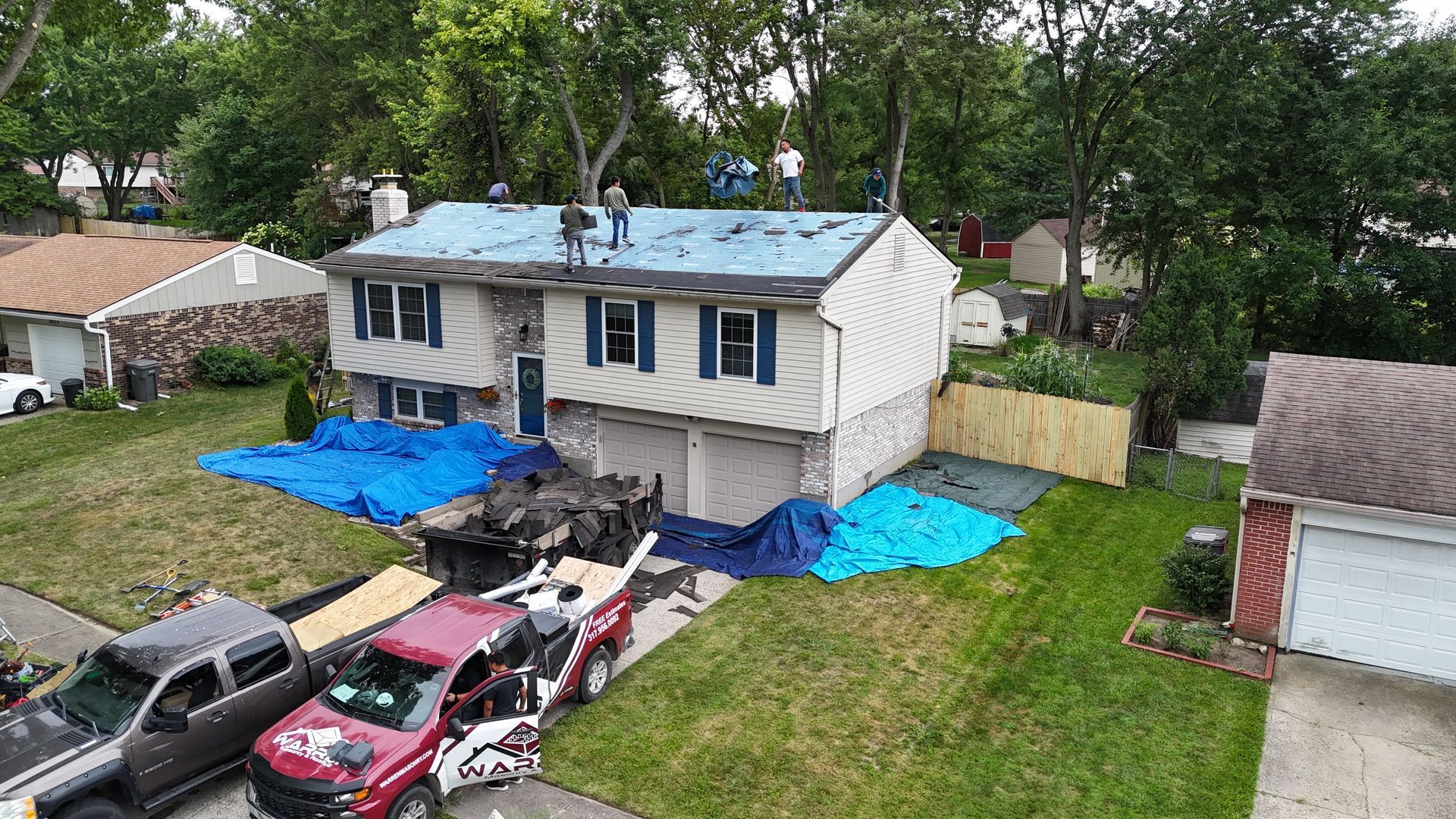 Workers on the roof of a two-story home during a residential roof replacement, with equipment and tarps on the lawn below.
