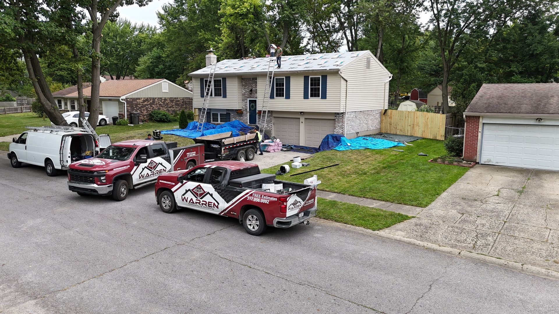 Roofers work on a house exterior with two red work trucks and a white van parked on the street.