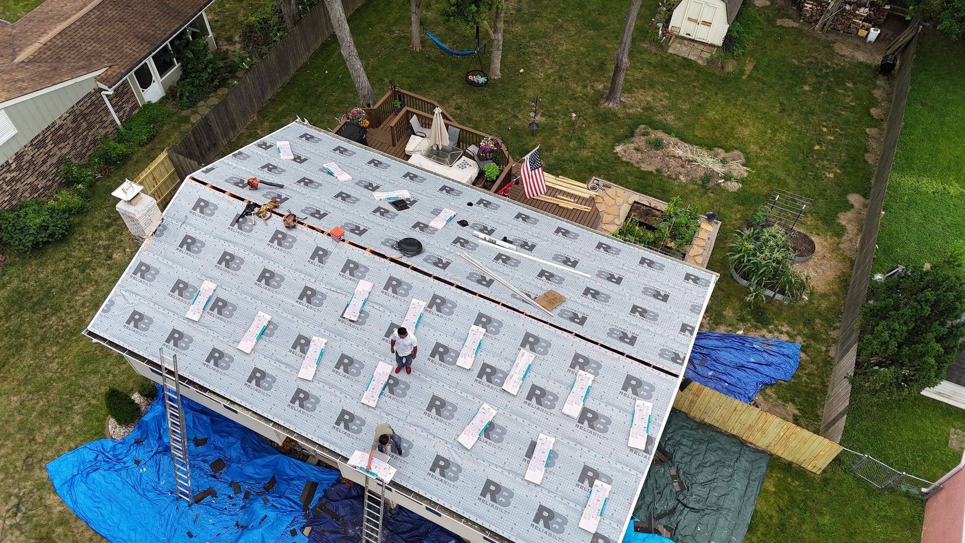 An aerial view shows workers installing synthetic underlayment on a gabled roof above a lawn with blue tarps.