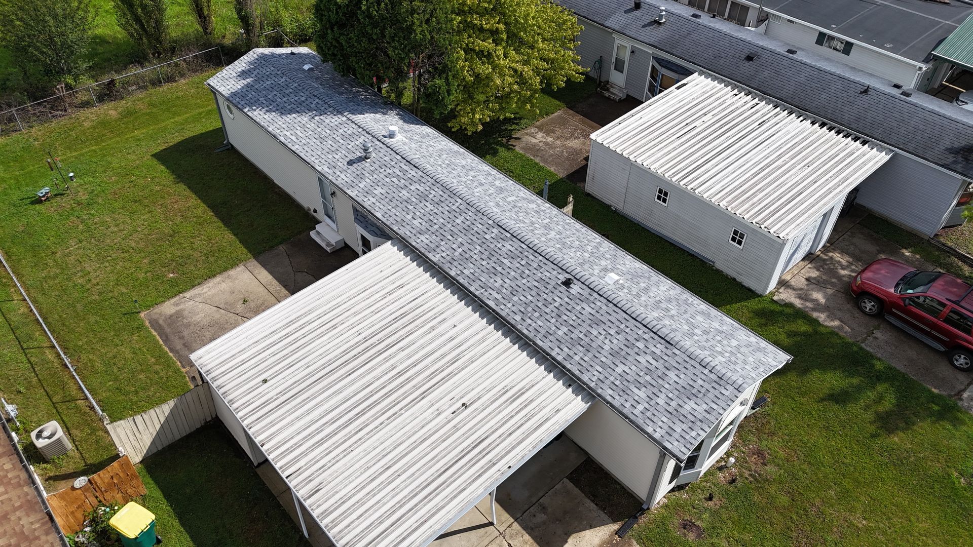 An aerial view of a long, white mobile home with a grey shingled roof, a front carport, and a separate small garage.