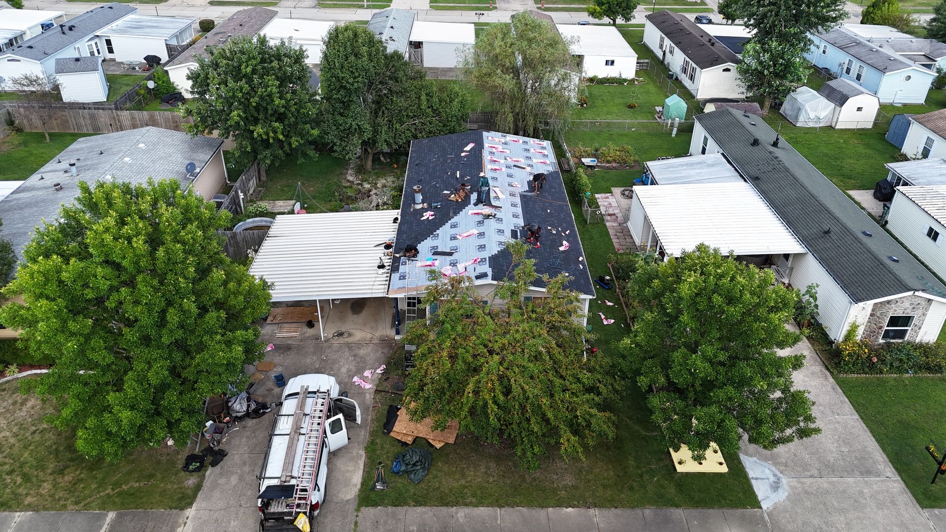 An aerial view of a house under renovation with a partially shingled roof, a work van in the driveway, and green trees.