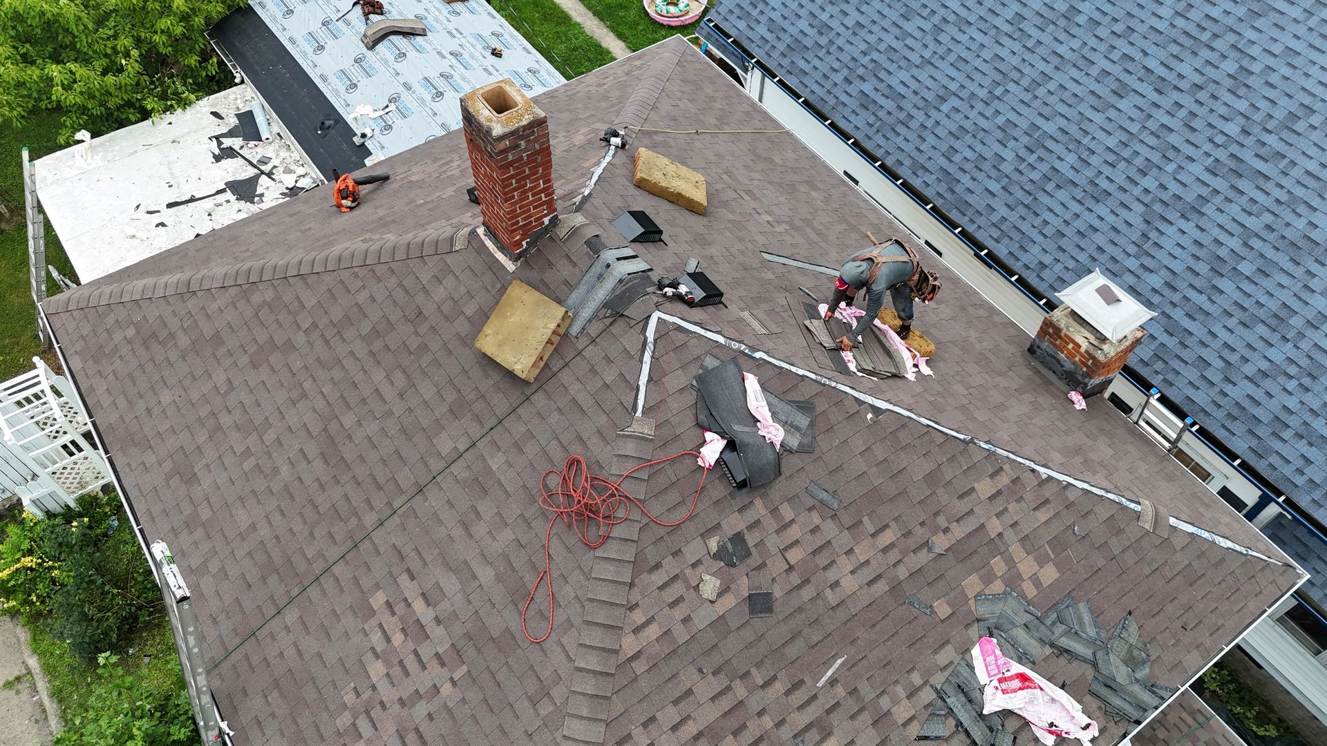 An aerial view shows workers installing brown asphalt shingles on a residential roof featuring chimneys and vents.