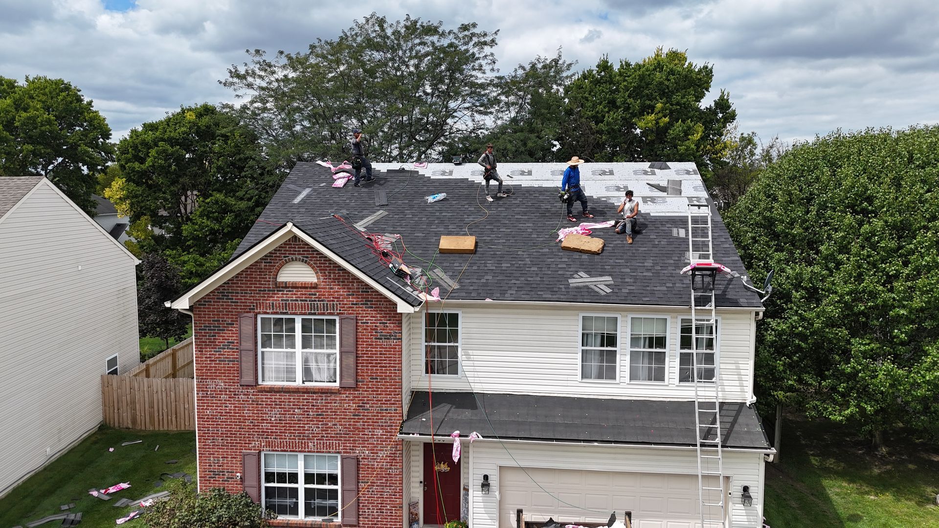 Workers install new black shingles on the roof of a two-story brick and white siding house during a sunny day.