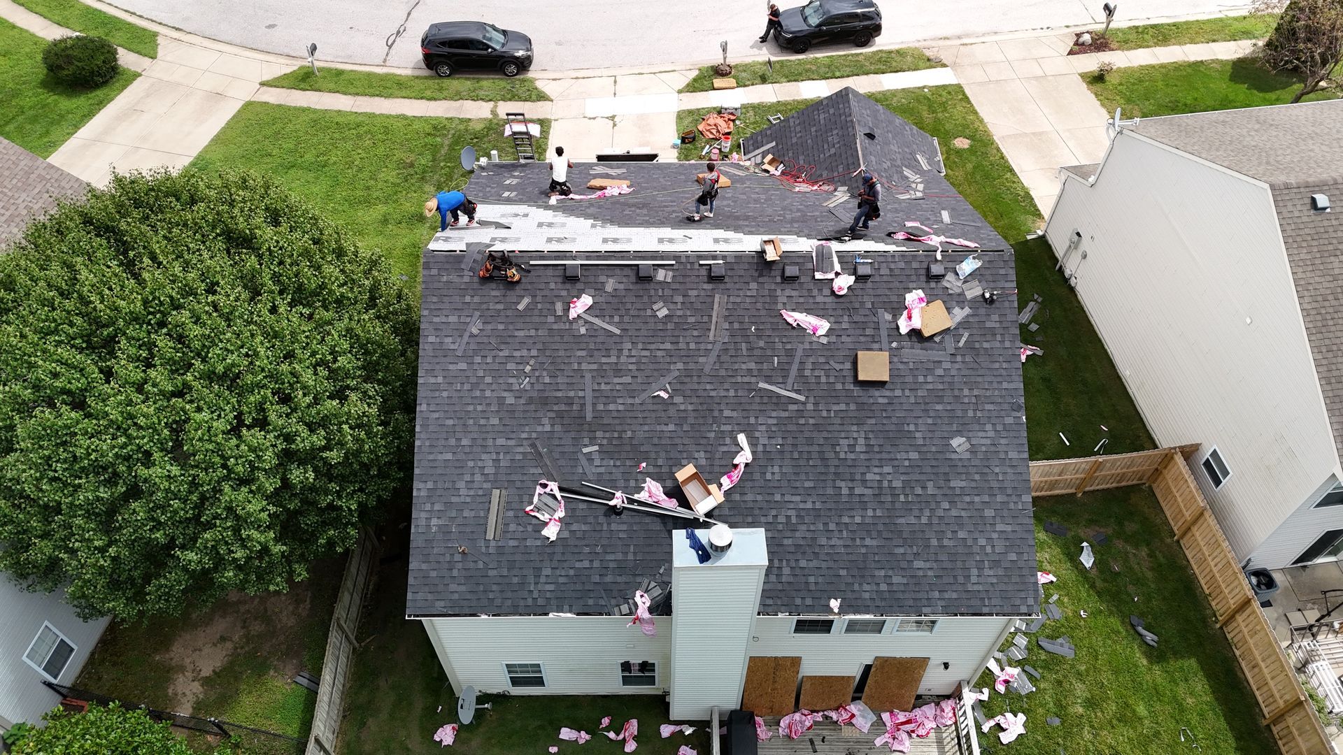 An aerial view shows workers on a residential roof removing shingles, with debris scattered across the roof and lawn.