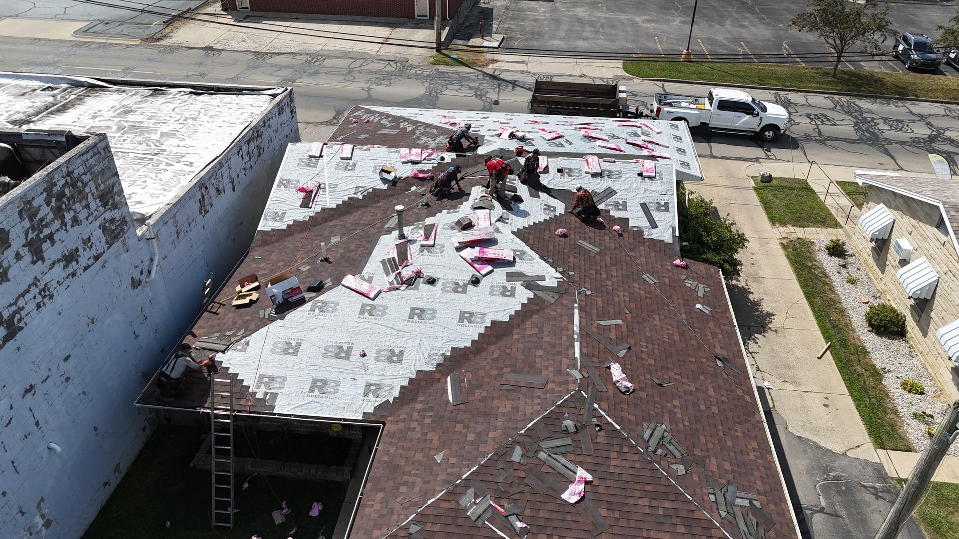 An aerial view of a building roof undergoing construction, with workers installing gray underlayment over brown shingles.