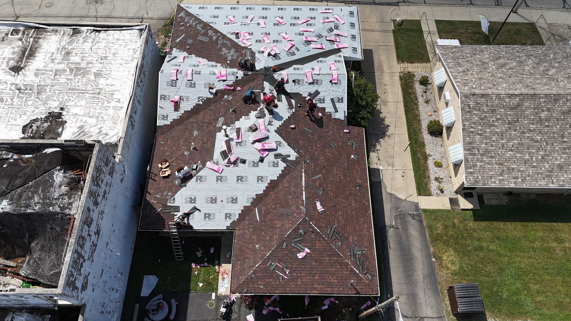 An aerial view shows workers replacing shingles on a house roof, with brown shingles and grey underlayment visible.