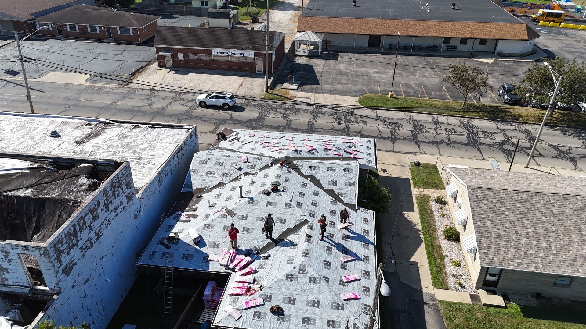 Aerial view of workers installing underlayment on a building roof, located on a street with nearby houses and parking lots.
