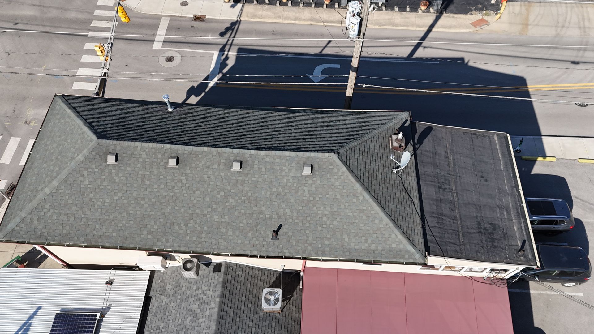 An aerial view of a building with a shingled, pitched roof and a flat roof section, next to a street intersection.