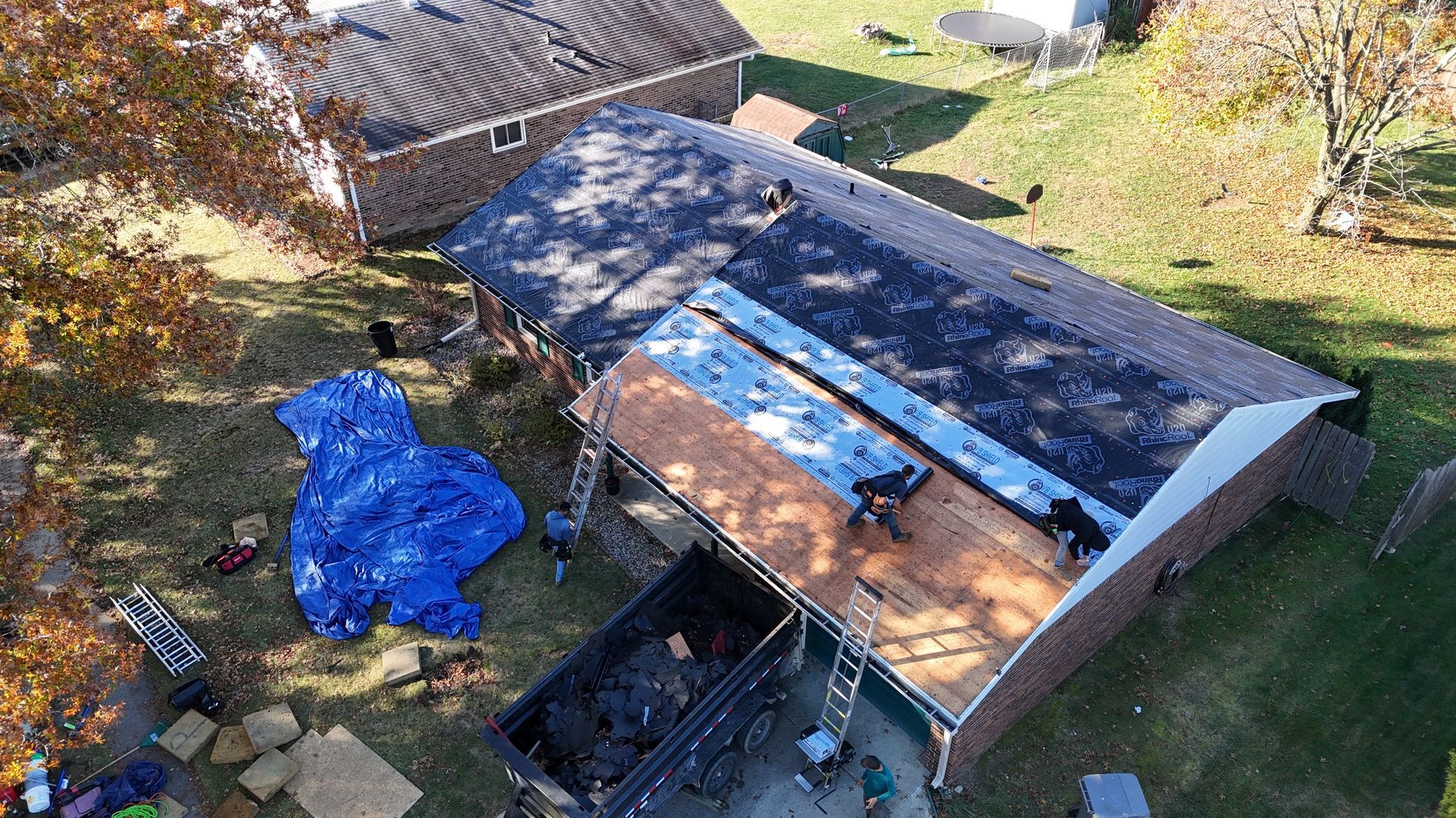 Aerial view of workers replacing a residential roof, with a dumpster and blue tarp on the ground below.