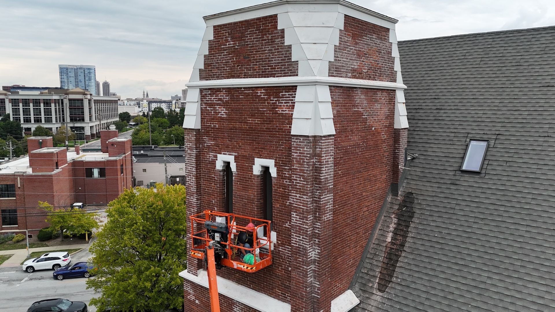 An orange aerial lift platform with workers repair a brick church tower next to a grey roof, with city buildings beyond.