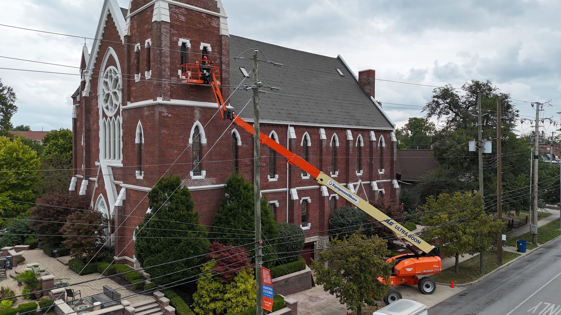An orange aerial lift reaches up to the side of a brick church, with a worker performing maintenance near a window.