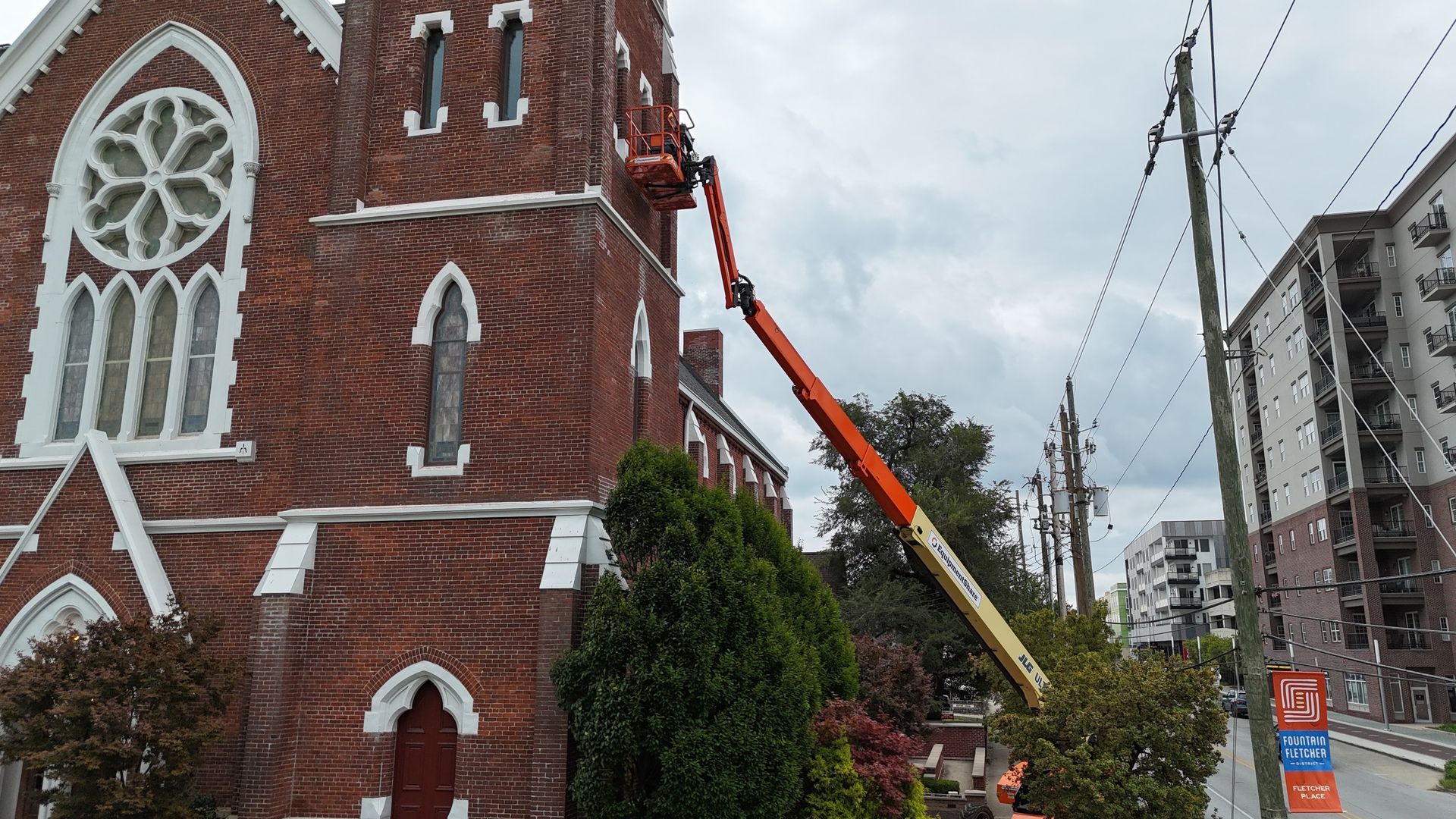 An orange boom lift is extended to the side of a historic red brick church for maintenance work.