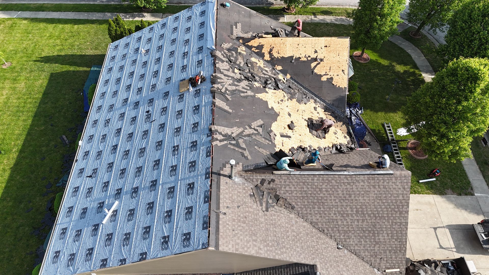 An aerial view shows roofers in the process of replacing shingles on a house, with sections in varying stages of repair.