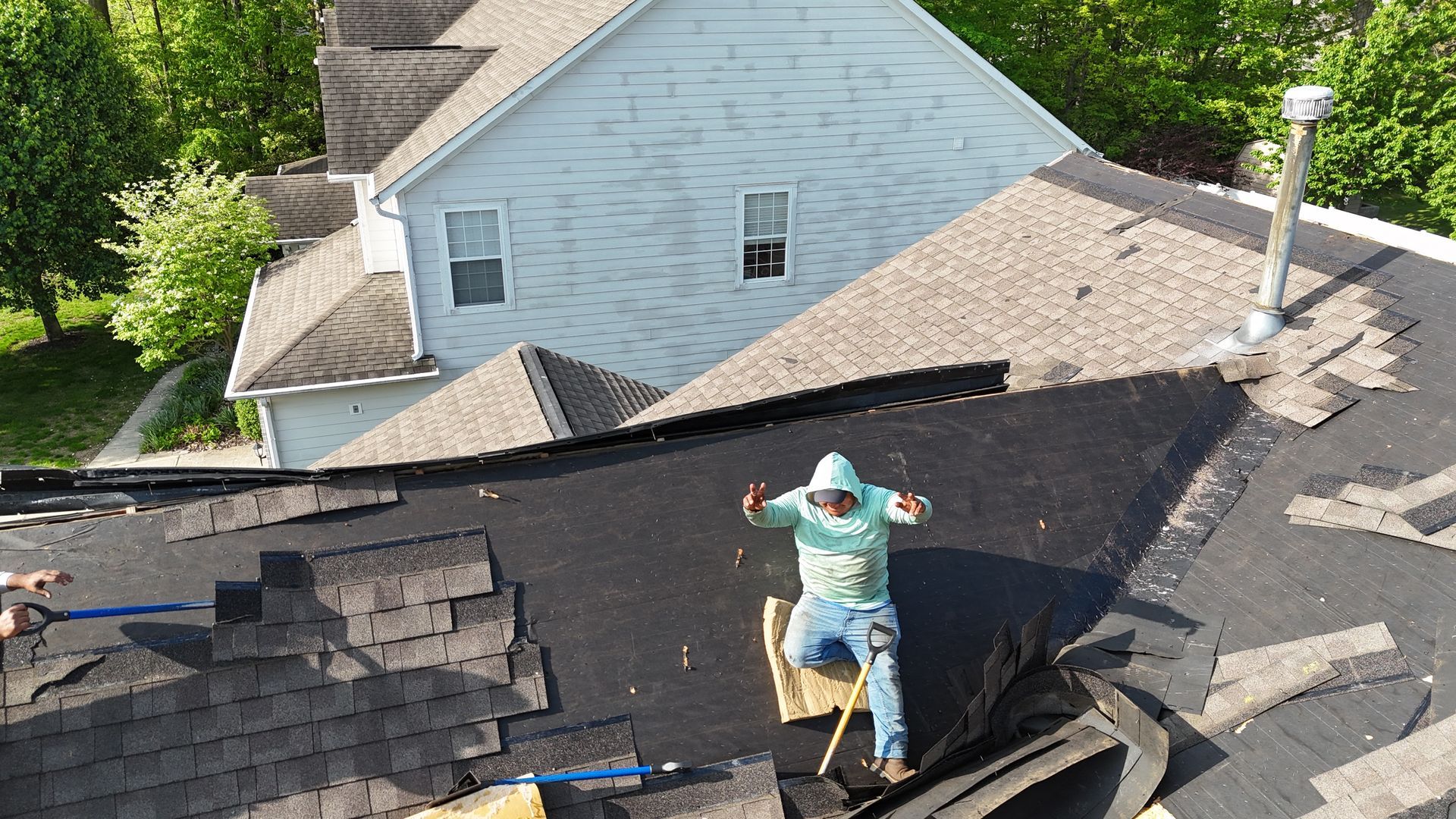 A person in a light blue hoodie sits on a partially stripped residential roof while working on home repairs.