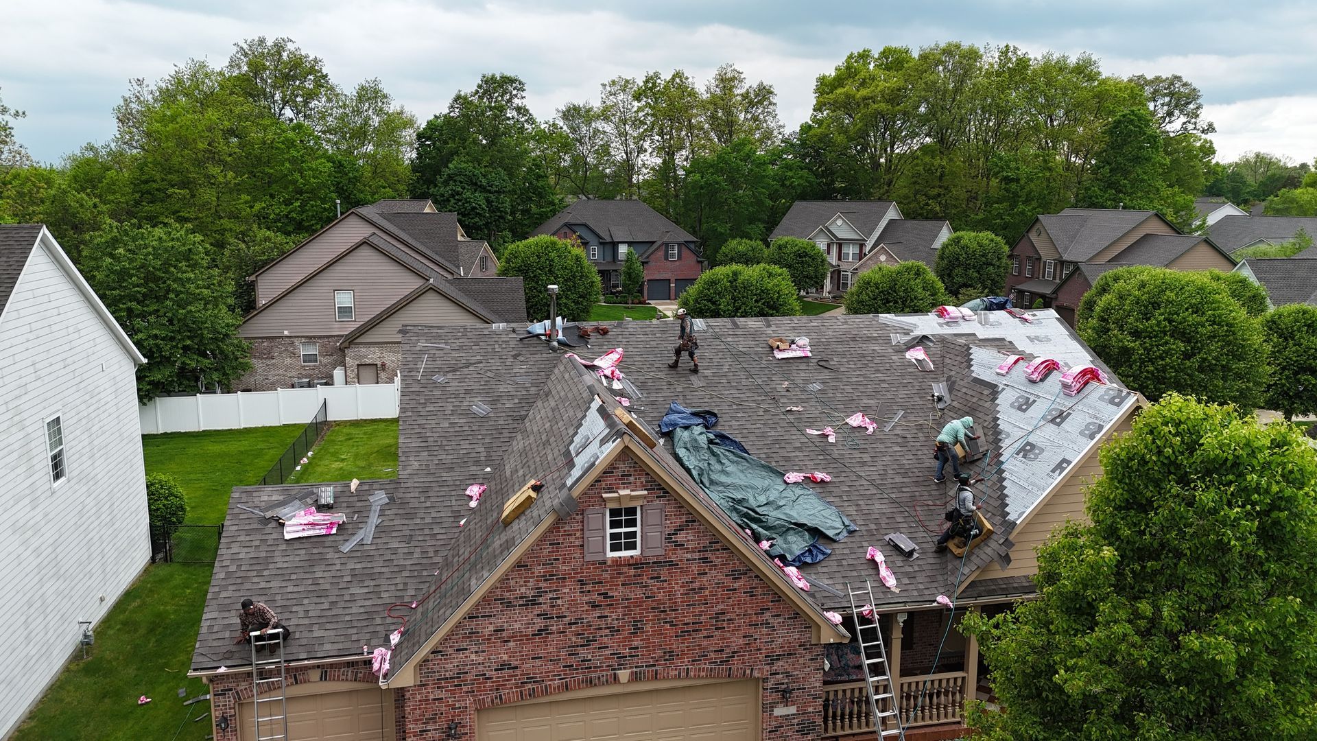 Construction workers performing a roof replacement on a brick suburban home on a cloudy day.